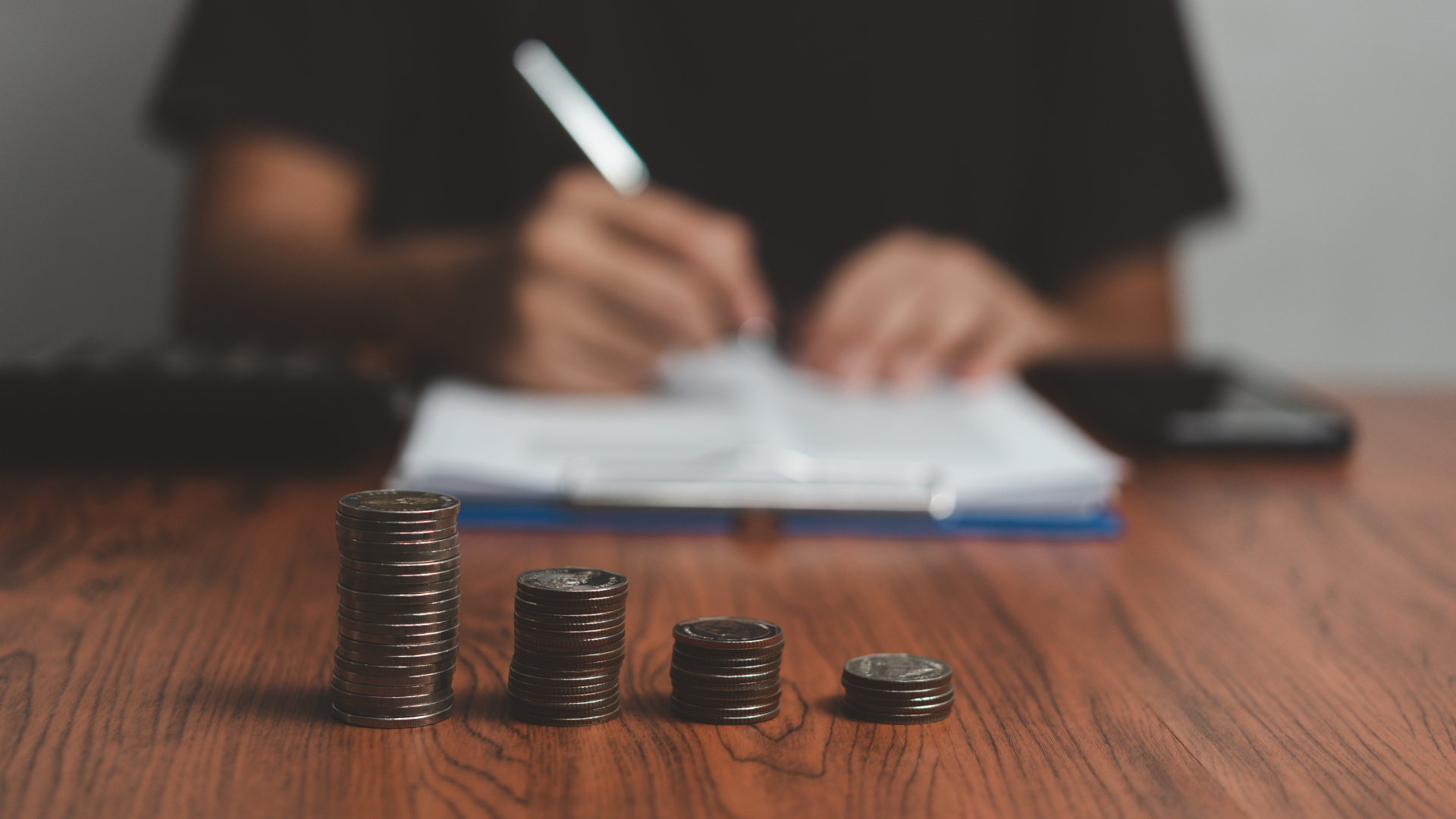 Pile di monete decrescenti su una scrivania di legno, dietro una persona compila moduli – stipendio dell'E-commerce Manager
